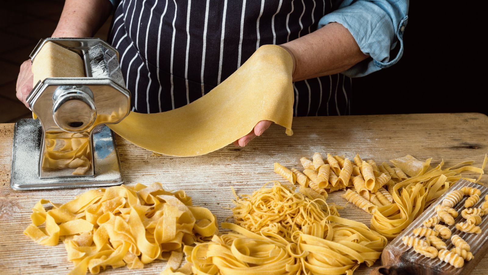 `Hands feeding fresh homemade dough through a pasta roller machine surrounded by freshly cut pasta noodles`