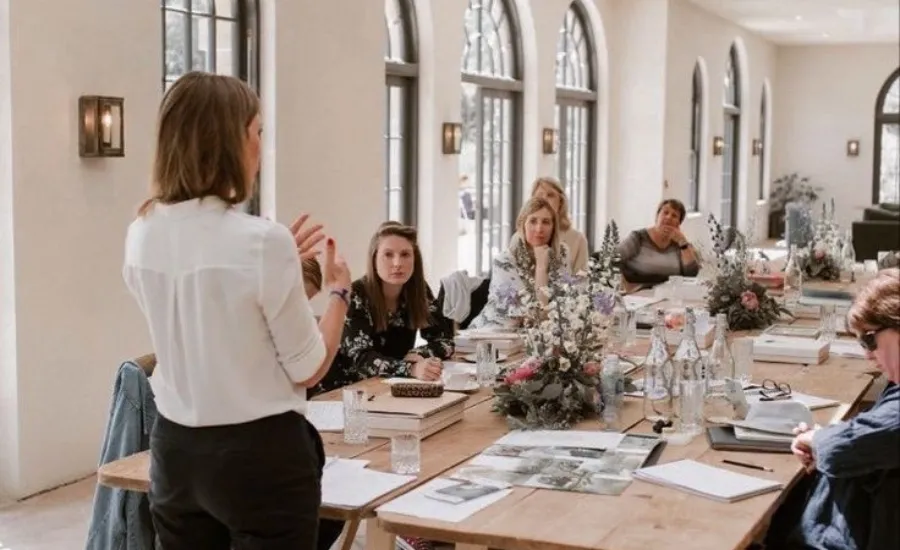 Woman speaking to an engaged audience seated around a floral decorated table in an airy modern conference room