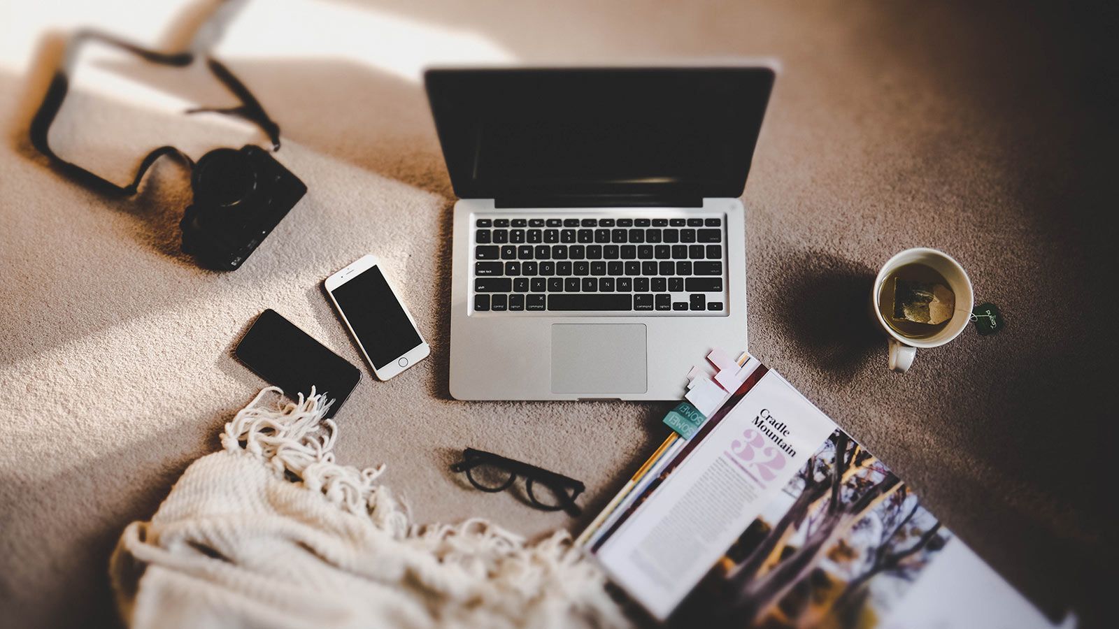 `High-angle flat lay of an open laptop, smartphones, vintage camera, magazine, and coffee mug arranged on a cozy textured blanket`
