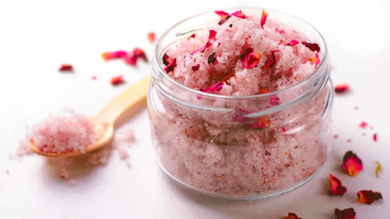 Glass jar of homemade pink salt and rose petal foot scrub with a wooden spoon