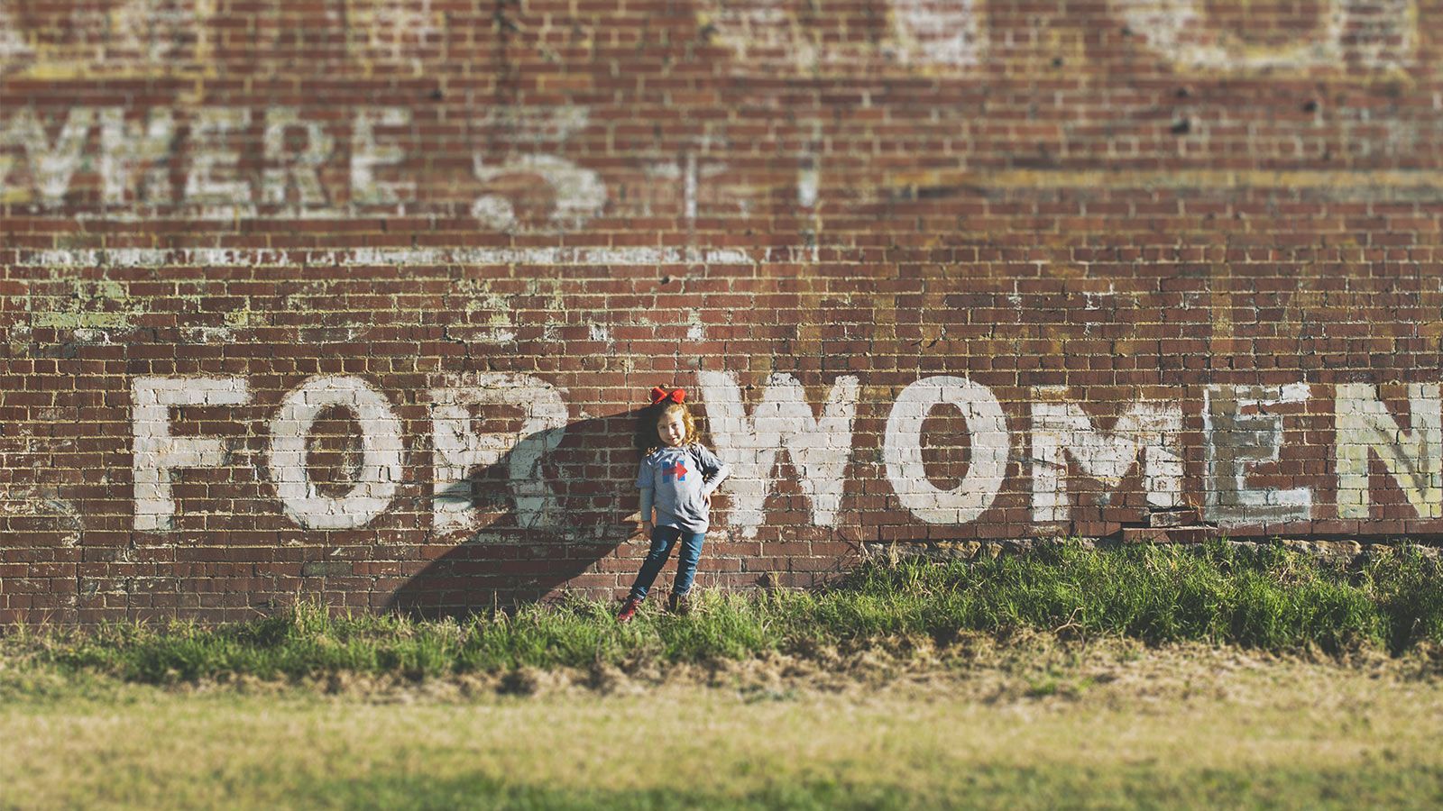 `Young woman standing in a grassy field in front of a weathered brick wall painted with "FOR WOMEN"`