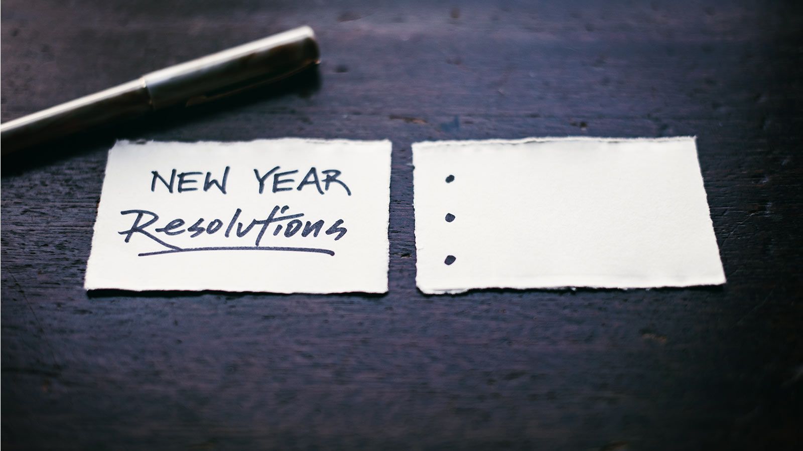 `Handwritten "New Year Resolutions" note beside a blank checklist on torn paper with a pen on a dark wooden surface`