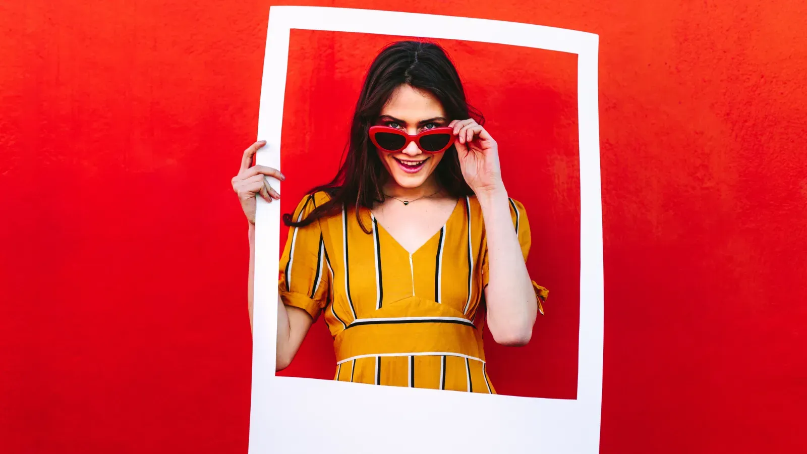 Young stylish woman holding an instant photo frame in front of herself against a red background