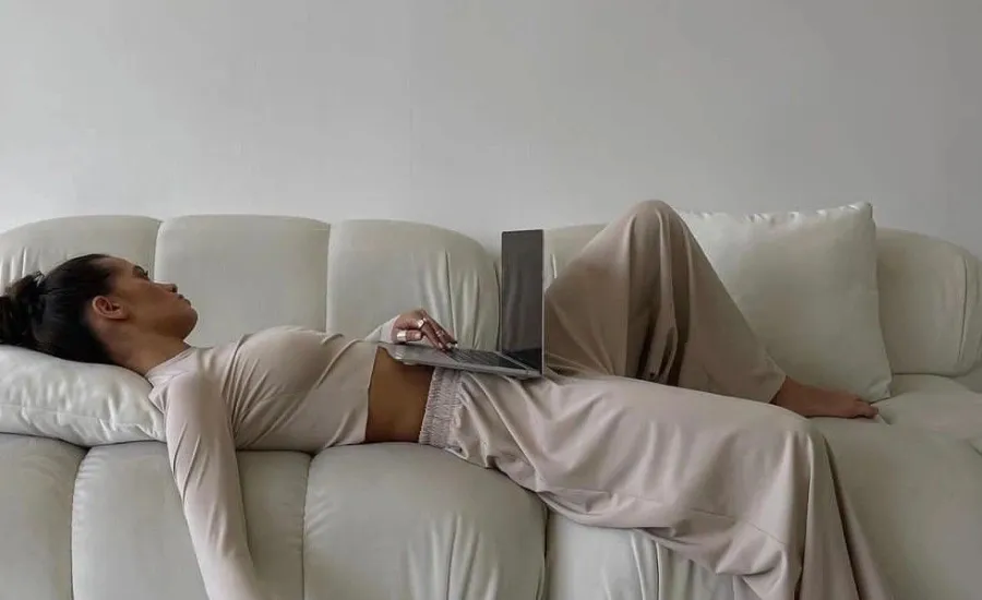Woman lying down on a white sofa working on an open laptop