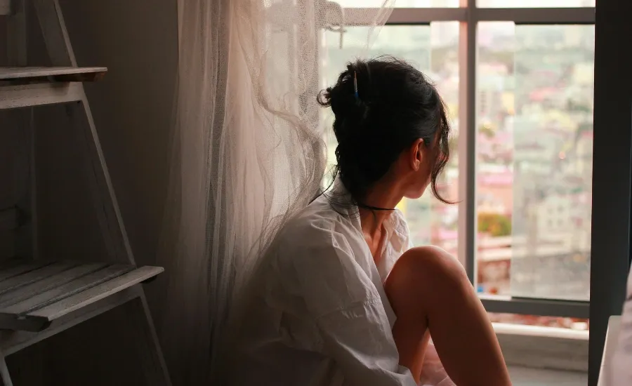 Pensive woman in a white shirt looking out of a large window overlooking the city