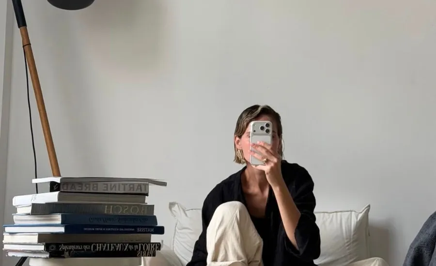 Woman taking a mirror selfie on bed next to a stack of art and lifestyle books