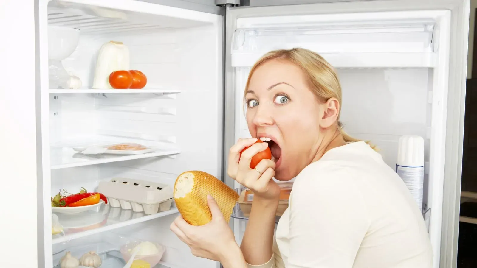 Hungry woman frantically eating food in front of an open refrigerator