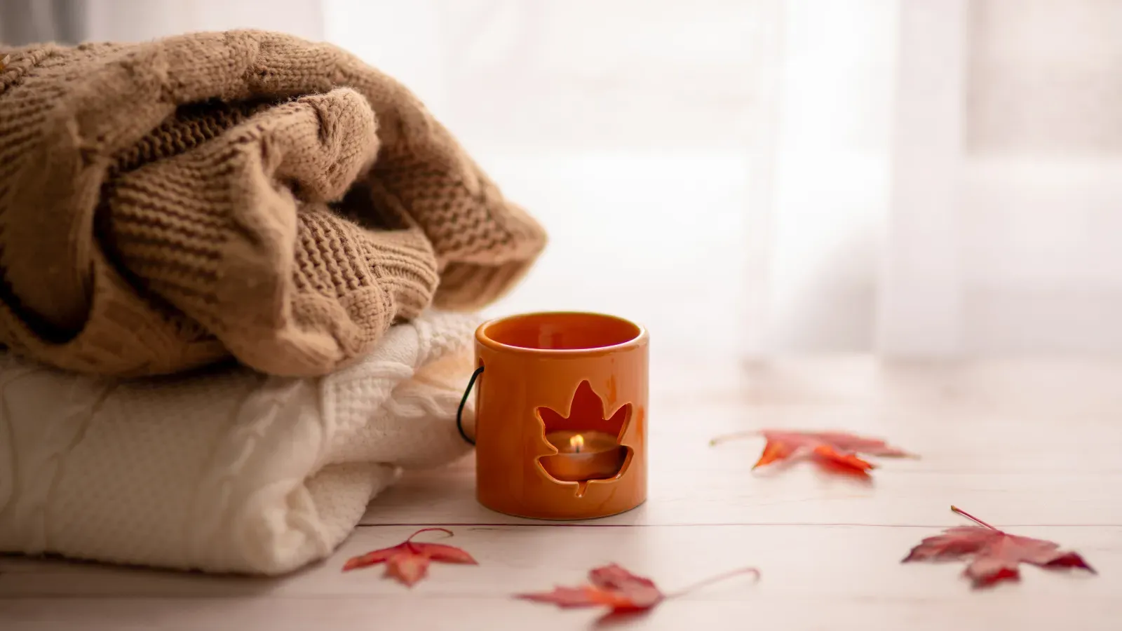 `Cozy folded knit sweaters next to a glowing autumn candle holder with scattered fall leaves by a window`