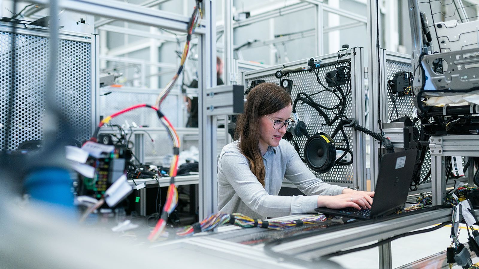 `Female engineer wearing glasses working on a laptop surrounded by complex wiring and hardware in a tech robotics laboratory`